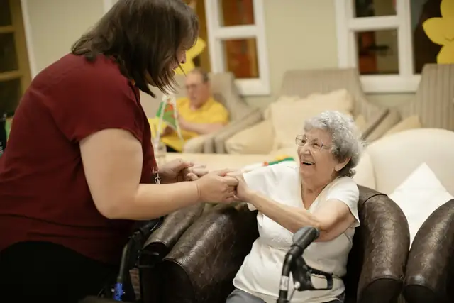 A carer holding hands with a smiling older woman in an armchair