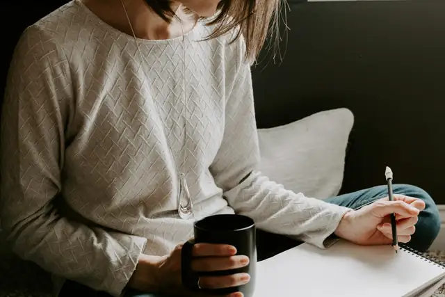 A woman writing in a notebook at a table, a coffee cup beside her