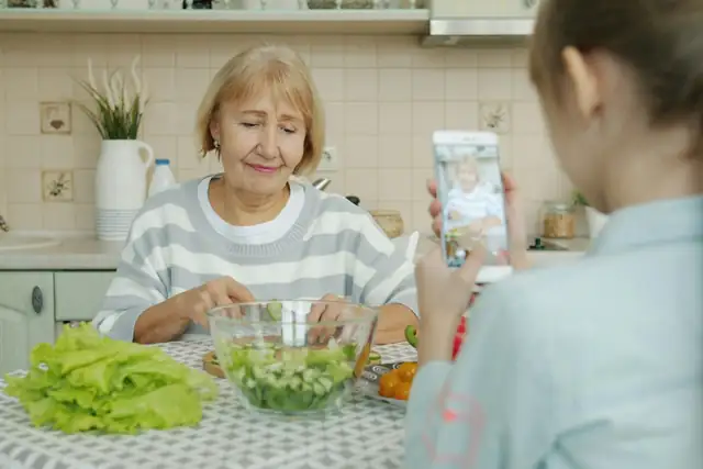 A grandmother making a salad while her granddaughter films her on a phone