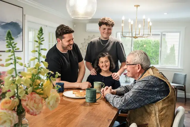 A family gathered around a wooden table with mugs and pastries, talking with an older man